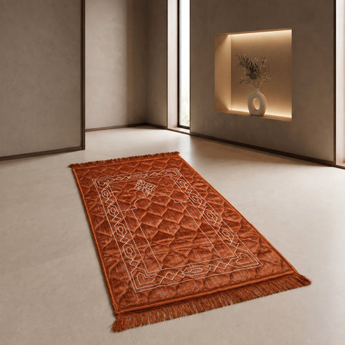 Rust quilted velvet prayer mat (janamaz) with embroidered geometric knot border and lantern, laid flat in a bright room